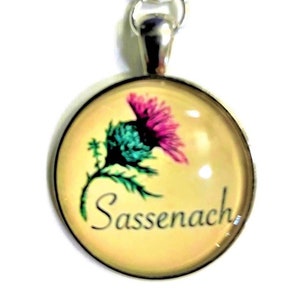 May include: A silver necklace with a round glass pendant featuring a pink and green thistle flower with the word "Sassenach" written in black script on a yellow background.