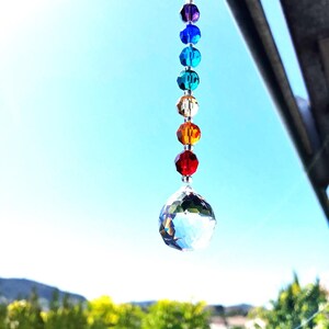 May include: A crystal suncatcher with a clear crystal ball and a string of rainbow colored beads. The suncatcher is hanging from a window.