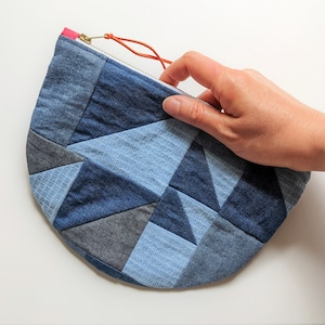 May include: A blue denim patchwork zippered pouch with a red zipper pull. The pouch is made of different shades of blue denim fabric and is shaped like a half-circle.