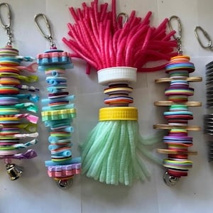 May include: Five colourful bird toys with different textures and shapes. Each toy has a metal ring for hanging and features a variety of colours, including pink, green, blue, yellow, and purple.