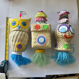 May include: Three colorful woven bird toys with hanging tassels. The toys are shaped like an owl, a box, and a head. The owl has yellow eyes and a pink beak. The box has a red, green, and blue circle pattern. The head has blue eyes and a red smile.