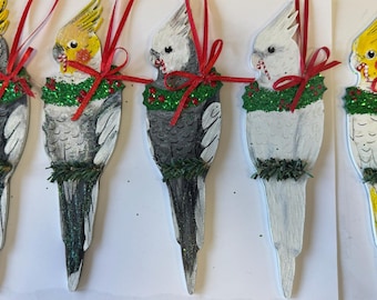 May include: Five wooden Christmas ornaments shaped like cockatiels. Each ornament is painted with a different color scheme, including gray, white, and yellow. The ornaments are decorated with a green and red holly wreath and a red ribbon.