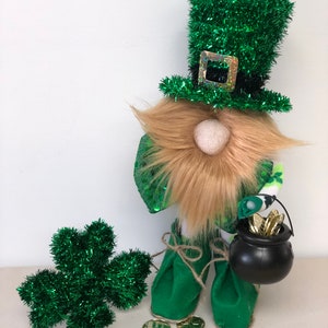 May include: A gnome figurine wearing a green tinsel top hat with a holographic buckle, a tan faux fur beard, and green fabric boots. The gnome holds a small black plastic cauldron filled with gold coins, with additional gold coins and a green tinsel shamrock.