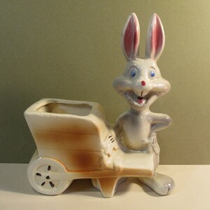 May include: A white ceramic planter shaped like a wheelbarrow with a cartoon rabbit pushing it. The rabbit has blue eyes, a red nose, and a wide smile. The planter is glazed in brown and white.