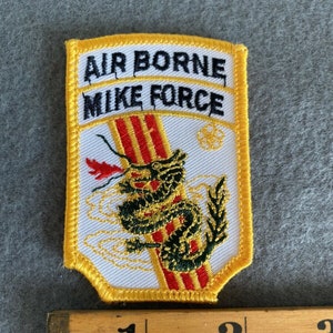 Pode incluir: Patch bordado com o texto "AIR BORNE MIKE FORCE" em preto sobre fundo branco. Uma borda amarela contorna o patch. Um desenho de dragão em verde, vermelho e dourado é apresentado no centro. Uma régua de madeira está ao lado do patch.