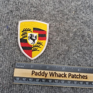 May include: Embroidered Porsche shield patch with a yellow, red, black, and white design. The patch features the word "PORSCHE" at the top and a black horse in the center. A wooden ruler with the text "Paddy Whack Patches" is below the patch.