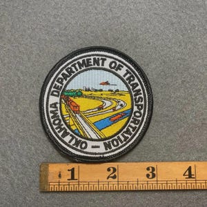 May include: Round patch featuring the Oklahoma Department of Transportation logo. The patch has a black border with white text. The center depicts a landscape with a train, road, and airplane. A wooden ruler is below the patch.