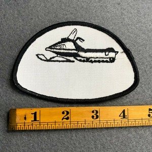 May include: Embroidered patch featuring a black and white snowmobile design on a white background. The patch has a black border and is approximately 4 inches long. Ideal for jackets, bags, or other fabric items.