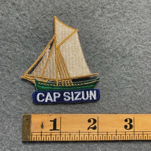 May include: Embroidered patch featuring a sailboat with beige sails and gold rigging, atop a green and blue base with the text "CAP SIZUN". The patch is approximately 2 inches long.