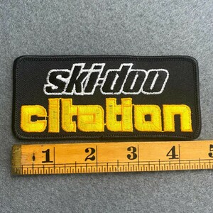 May include: Black rectangular embroidered patch with the words "ski-doo citation" in white and yellow. The patch is approximately 4 inches long. A wooden ruler is placed below the patch.