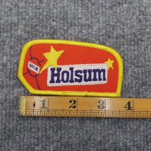 May include: A rectangular patch with a yellow border and a red interior. The patch features the word "Holsom" in white and blue lettering, along with yellow stars and a small map graphic. A ruler is placed below the patch.