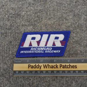 May include: A blue embroidered patch with the text "RIR" in white, above "RICHMOND INTERNATIONAL RACEWAY". The patch has red stripes and is rectangular. A wooden ruler with inches is below the patch.