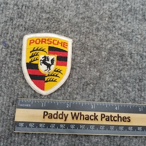 May include: Embroidered Porsche badge featuring the brand's crest in yellow, black, red, and white. The badge is shield-shaped and has the Porsche name in red. A wooden ruler with inch measurements and the text "Paddy Whack Patches" is also visible.