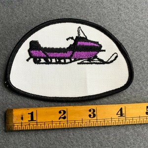 May include: Embroidered patch featuring a purple and black snowmobile design on a white background. The patch has a black border and is approximately 3 inches long. Ideal for jackets, bags, or other fabric items.
