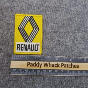 May include: A yellow and black Renault patch with the brand name in white text. The patch features a diamond-shaped logo with black and white stripes. The patch is rectangular and has a stitched border. A wooden ruler is below the patch.