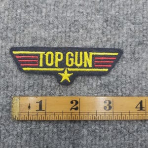 Może przedstawiać: Prostokątna naszywka Top Gun z żółtym napisem i gwiazdą, czerwonymi paskami i czarną obwódką. Naszywka ma około 7.6 cm szerokości. Naszywka znajduje się na szarej powierzchni.