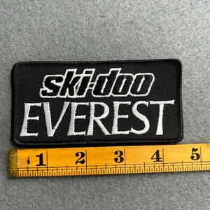 May include: Black rectangular patch with the text "ski-doo EVEREST" in white. The patch is approximately 4 inches long. The patch is on a gray surface.