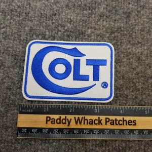 May include: A rectangular patch with a white background and a blue border. The word "COLT" is embroidered in blue, with a stylized "C" and "T". A wooden ruler with inch markings and the text "Paddy Whack Patches" is below the patch.