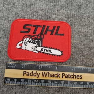 Puede incluir: Un parche rectangular rojo con la palabra "STIHL" en negro y una ilustración de motosierra blanca y negra. La motosierra tiene la palabra "STIHL" en la hoja. Una regla con pulgadas está en la imagen.