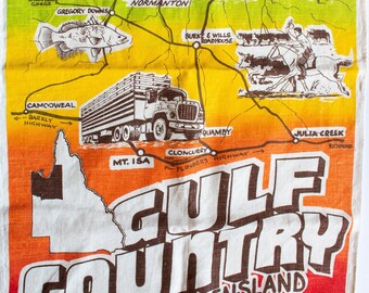 Op de afbeelding: Een vintage-stijl kaart van Gulf Country, Noord-Queensland, Australië. De kaart is gedrukt op een witte linnen theedoek en heeft een kleurrijke illustratie van de bezienswaardigheden van de regio, waaronder de stad Normanton, de Gregory Downs en het Burke & Wills Roadhouse. De kaart bevat ook een krokodil, een vrachtwagen en een boot.