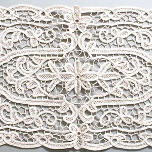 May include: Cream-colored, rectangular lace doily with a floral and scrollwork design. The delicate lace features a repeating pattern of flowers and swirling vines, creating an intricate and elegant textile.