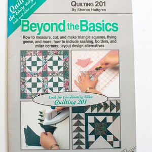 Op de afbeelding: Een boek getiteld "Quilting 201 Beyond the Basics" door Sharon Hultgren. De omslag toont quiltvoorbeelden, waaronder driehoekige vierkanten en vliegende ganzen. Het boek maakt deel uit van een "Quilt the easy way"-serie en bevat een bijbehorende video. De omslag is grijs en blauwgroen.