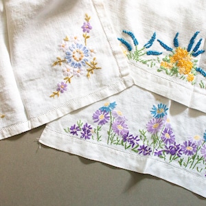 以下が含まれることがあります： 白のリネン生地に、青、黄、紫色の花の刺繍が施されています。 刺繍は、花と葉の細かい模様で、手作りの風合いを加えています。 生地は折りたたまれ、刺繍された縁が際立っています。