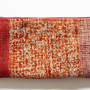 May include: A rectangular, zippered pouch with a textured fabric exterior. The pouch is divided into three sections of color: solid red, speckled orange and white, and a mix of red and brown with yellow threads. The zipper is red.