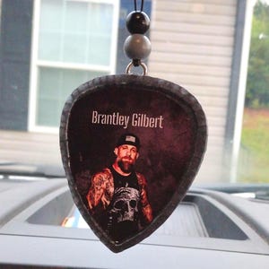 May include: A guitar pick-shaped car air freshener featuring a portrait of Brantley Gilbert. The image shows Gilbert wearing a black hat and skull tank top. The air freshener is suspended by a black cord with gray and black beads.