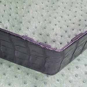 Puede incluir: Una cartera de cuero negro con un patr&oacute;n de cocodrilo. La cartera est&aacute; doblada por la mitad y est&aacute; sobre una superficie verde.