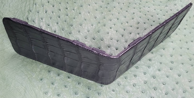 Puede incluir: Una cartera de cuero negro con un patr&oacute;n de cocodrilo. La cartera est&aacute; doblada por la mitad y est&aacute; sobre una superficie verde.