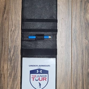 Puede incluir: Porta tarjetas de puntuaci&oacute;n de golf de cuero negro con un l&aacute;piz azul en el interior. El porta tarjetas tiene una tarjeta blanca con el texto "UNDER ARMOUR", "THE JUNIOR TOUR", "UAGOLFTOUR.COM", "NATIONAL CHAMPIONSHIP", "February 12th-15th, 2021" y "Tournament Yardage Book".