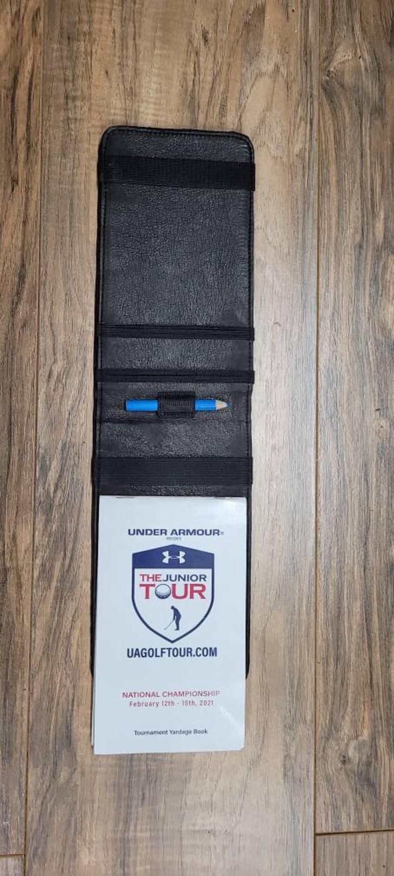Puede incluir: Porta tarjetas de puntuaci&oacute;n de golf de cuero negro con un l&aacute;piz azul en el interior. El porta tarjetas tiene una tarjeta blanca con el texto "UNDER ARMOUR", "THE JUNIOR TOUR", "UAGOLFTOUR.COM", "NATIONAL CHAMPIONSHIP", "February 12th-15th, 2021" y "Tournament Yardage Book".