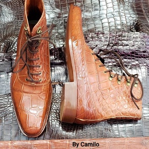 May include: A pair of brown alligator leather boots with laces. The boots are on a brown alligator skin background. The text "By Camilo Genuine alligator" is visible at the bottom of the image.