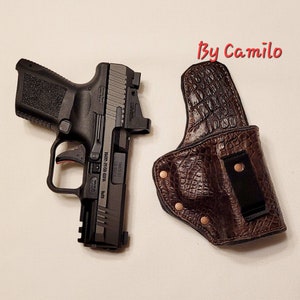 May include: A black handgun with a brown leather holster. The holster has a crocodile pattern and a black clip. The handgun has a red safety switch and a black sight.