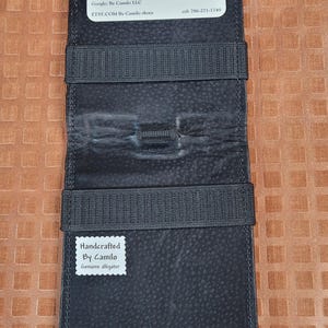 Puede incluir: Cartera de cuero negro con cierre de correa. La cartera tiene un porta tarjetas de visita y una etiqueta que dice "Handcrafted By Camilo Genuine alligator".