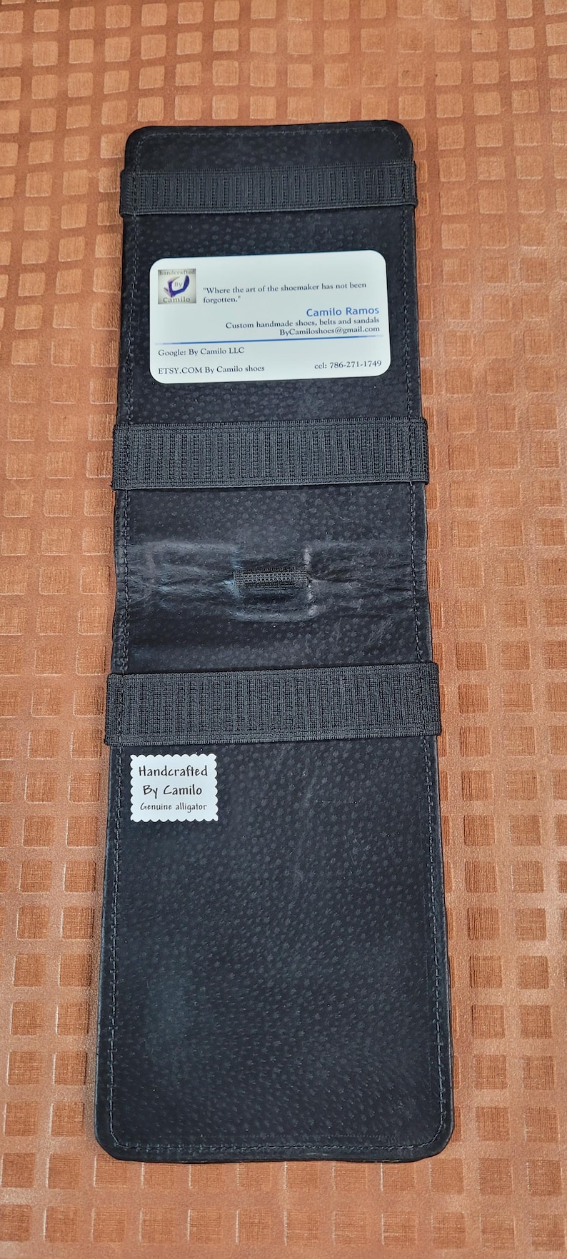 Puede incluir: Cartera de cuero negro con cierre de correa. La cartera tiene un porta tarjetas de visita y una etiqueta que dice "Handcrafted By Camilo Genuine alligator".