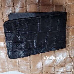 Puede incluir: Cartera de cuero negro con un patr&oacute;n de cocodrilo y el texto "Genuine Alligator" grabado en la parte delantera. La cartera est&aacute; sobre un fondo marr&oacute;n.