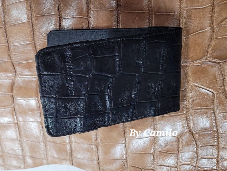 Puede incluir: Cartera de cuero negro con un patr&oacute;n de cocodrilo y el texto "Genuine Alligator" grabado en la parte delantera. La cartera est&aacute; sobre un fondo marr&oacute;n.