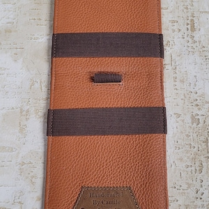 Puede incluir: Una cartera de cuero marr&oacute;n con bandas el&aacute;sticas marrones y un parche de cuero marr&oacute;n que dice "Handcrafted By Camilo Genuine alligator".
