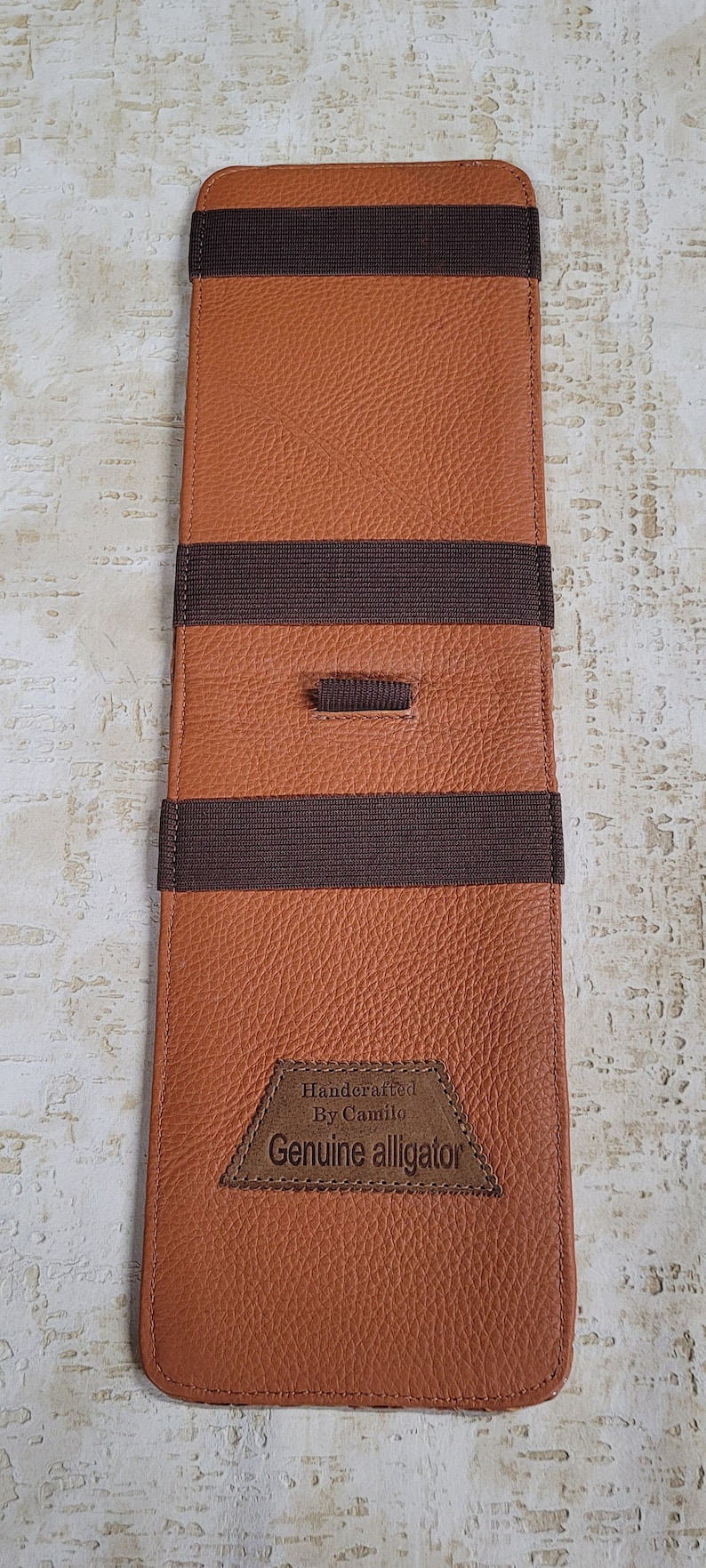 Puede incluir: Una cartera de cuero marr&oacute;n con bandas el&aacute;sticas marrones y un parche de cuero marr&oacute;n que dice "Handcrafted By Camilo Genuine alligator".