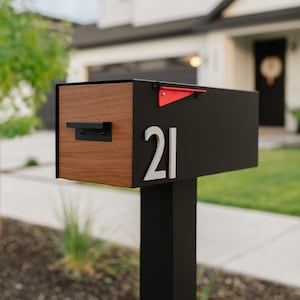 May include: A modern mailbox with a black body and a wooden front panel. The mailbox features the number "21" in silver and a red flag. It is mounted on a black post, suitable for residential use.