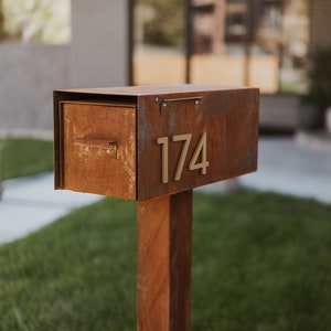 May include: A rusty metal mailbox with a number "174" attached to the front. The mailbox is mounted on a rusty metal post.