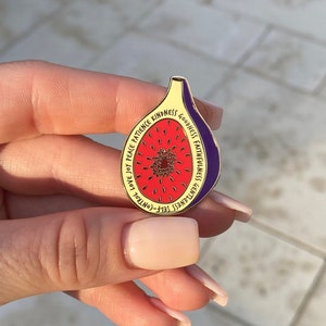 Op de afbeelding: Gouden speld in de vorm van een vijg met een rood en wit interieur. De speld heeft de woorden "Zelfbeheersing, Zachtaardigheid, Getrouwheid, Goedheid, Liefde, Geduld, Vrede, Vreugde, Liefde" rond de rand.