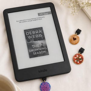May include: A black Kindle e-reader displaying a book cover and text, with three decorative charms. The charms include a cookie with a heart, a pizza, and a blue charm with the text "it is well with my soul."