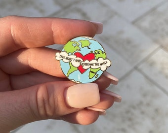 May include: A gold-colored enamel pin featuring a blue and green globe with a red heart in the center. The globe is surrounded by a white cloud with the text "Take heart I have overcome the world".
