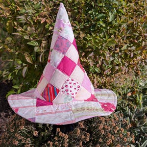 May include: A conical hat constructed from a patchwork of various pink and white fabric squares. The hat features a wide, circular brim and a pointed crown, with visible stitching connecting the diverse patterned textile pieces throughout.