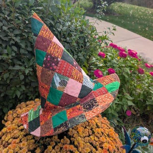 May include: A pointed hat constructed from a patchwork of various fabric squares in shades of green, brown, orange, and pink. The hat features a wide, slightly curved brim and a tall, conical crown, all stitched together with visible seams.