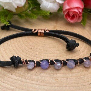 May include: Black leather bracelet with copper wire accents and lavender-colored beads. The bracelet features a sliding knot closure for adjustable sizing. The beads are a mix of round and faceted shapes, adding texture to the design.
