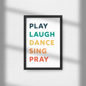 Könnte beinhalten: Ein schwarzer gerahmter Druck mit den Worten "PLAY", "LAUGH", "DANCE", "SING" und "PRAY" in bunten Schriftzügen.